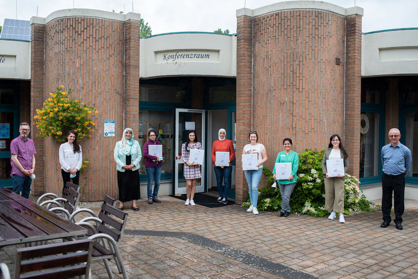 10 Personen stehen vor einem Gebäude und halten Unterlagen in der Hand. Vor dem Gebäude steht die Aufschrift 'Konferenzraum'.