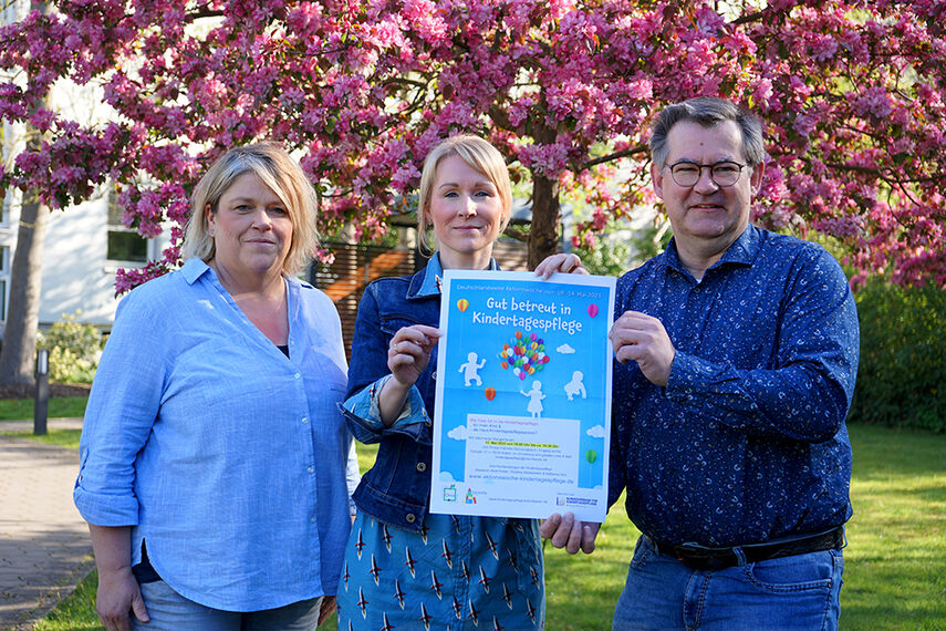 3 Personen stehen im Garten und halten ein Plakat zum Thema 'Gut betreut in Kindertagespfege'